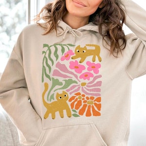 May include: A beige hoodie with a graphic print of two orange cats and pink and orange flowers. The text "Sand" is printed on the bottom of the hoodie.