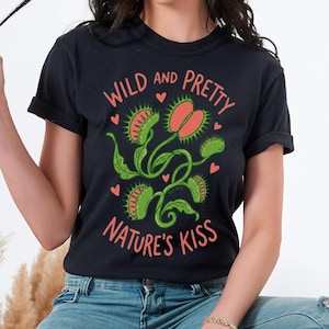 Puede incluir: Camiseta negra con un gráfico de atrapamoscas y el texto "WILD AND PRETTY NATURE'S KISS" en coral y verde. El diseño incluye formas de corazón. La camiseta la lleva una persona.