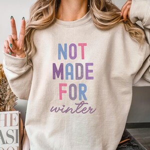 May include: A light beige sweatshirt with the text "NOT MADE FOR winter" in colorful letters. The text is in a distressed font style.
