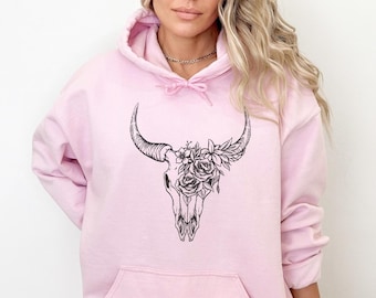 Può includere: Una felpa con cappuccio rosa chiaro con tasca a marsupio e cappuccio con coulisse. La parte anteriore presenta una grafica in bianco e nero di un teschio di toro adornato con fiori. La felpa è realizzata in un materiale morbido e confortevole.