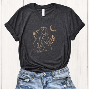 K&ouml;nnte beinhalten: Dunkelgraues Heather-T-Shirt mit einem goldenen Linienkunst-Design einer sitzenden Figur, Sternen und einem Halbmond. Das Design beinhaltet florale Elemente. Das Shirt wird mit Jeansshorts kombiniert.
