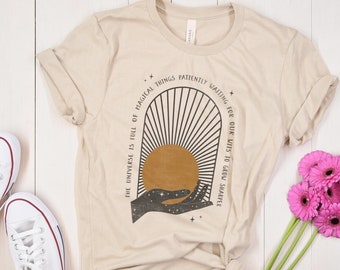 May include: A beige t-shirt with a graphic design of a sun rising over a hand with the text "The universe is full of magical things patiently waiting for our wits to grow sharper".