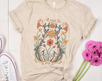 May include: A beige t-shirt with a floral design featuring a sun face, flowers, and mushrooms. The design is in a vintage style with a green, orange, and yellow color scheme.