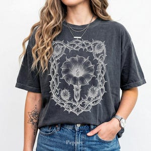 Puede incluir: Camiseta gris oscuro con un dise&ntilde;o floral blanco detallado dentro de un marco espinoso. El dise&ntilde;o incluye una flor grande y elementos similares a cardos. La camiseta es de corte holgado, con un lavado vintage.