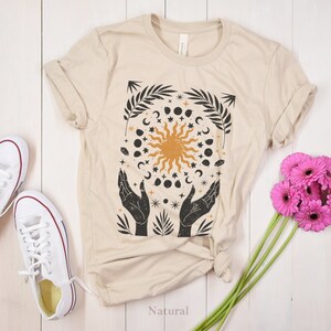 Könnte beinhalten: Ein beiges T-Shirt mit einem grafischen Druck einer Sonne, eines Mondes und Sternen, die von Händen umgeben sind. Die Grafik ist in Schwarzweiß mit einer gelben Sonne.