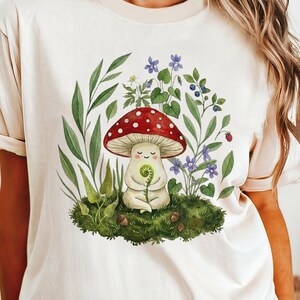 May include: Cream-colored t-shirt featuring a whimsical illustration of a mushroom character with a red and white spotted cap, surrounded by greenery and flowers. The mushroom is holding a green sprout. The design is centered on the shirt.