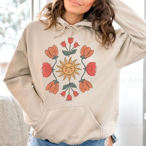 May include: A beige hoodie with a graphic print of a sun with a face and flowers in shades of red, orange, and green. The text "Sand" is printed on the bottom of the hoodie.