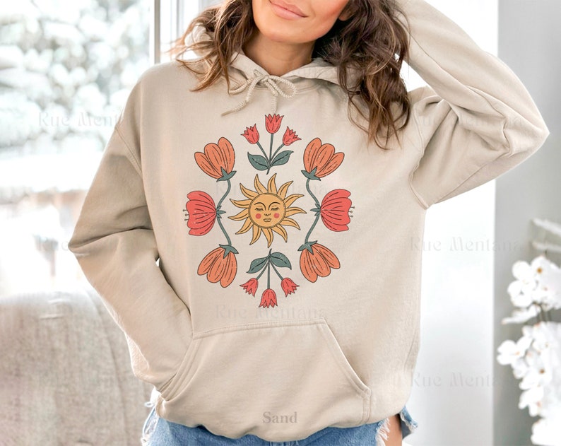 May include: A beige hoodie with a graphic print of a sun with a face and flowers in shades of red, orange, and green. The text "Sand" is printed on the bottom of the hoodie.