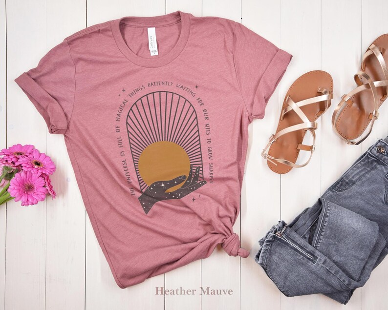 May include: A heather mauve t-shirt with a graphic design of a hand reaching for a sun with the text "The universe is full of magical things patiently waiting for our wits to grow sharper".
