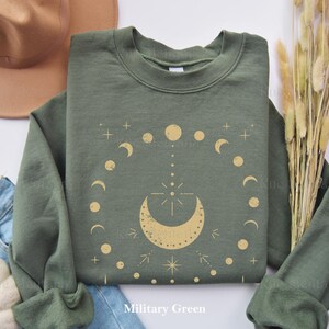 以下が含まれることがあります： 軍緑色のスウェットシャツに、金の月と星のデザインが施されています。デザインは、三日月の中に星が描かれており、その周りを小さな月と星が円形に囲んでいます。