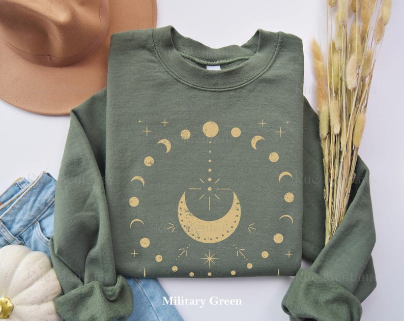 以下が含まれることがあります： 軍緑色のスウェットシャツに、金の月と星のデザインが施されています。デザインは、三日月の中に星が描かれており、その周りを小さな月と星が円形に囲んでいます。