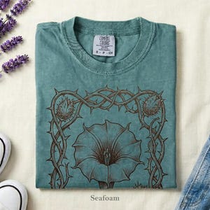 Puede incluir: Camiseta verde espuma de mar con un dise&ntilde;o floral marr&oacute;n y la palabra "Seafoam" en la parte inferior. La camiseta est&aacute; doblada y colocada sobre una superficie blanca junto a un par de zapatillas blancas, lavanda y vaqueros.