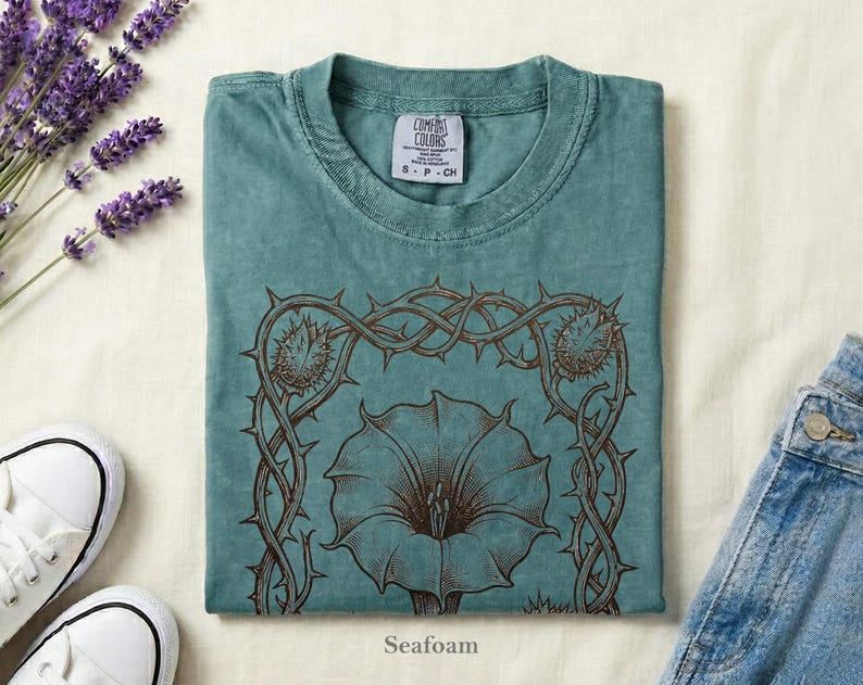Puede incluir: Camiseta verde espuma de mar con un dise&ntilde;o floral marr&oacute;n y la palabra "Seafoam" en la parte inferior. La camiseta est&aacute; doblada y colocada sobre una superficie blanca junto a un par de zapatillas blancas, lavanda y vaqueros.