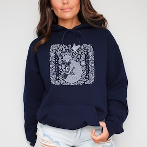 May include: Navy blue hoodie with a white fox design in a square floral frame. The fox is curled up, surrounded by detailed floral and bird illustrations. The hoodie has a drawstring hood and a front pocket.