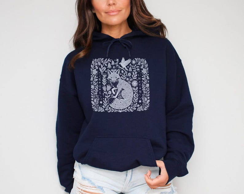 May include: Navy blue hoodie with a white fox design in a square floral frame. The fox is curled up, surrounded by detailed floral and bird illustrations. The hoodie has a drawstring hood and a front pocket.