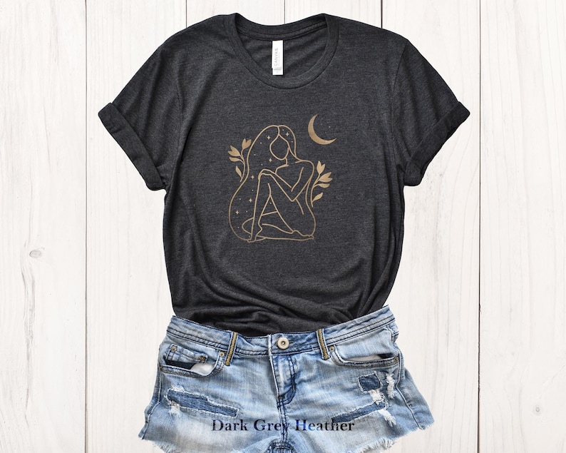 K&ouml;nnte beinhalten: Dunkelgraues Heather-T-Shirt mit einem goldenen Linienkunst-Design einer sitzenden Figur, Sternen und einem Halbmond. Das Design beinhaltet florale Elemente. Das Shirt wird mit Jeansshorts kombiniert.