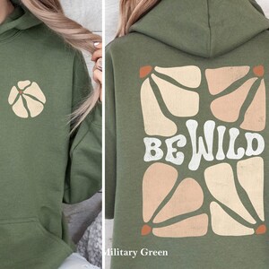 Op de afbeelding: Een groene hoodie met een witte "Be Wild" afbeelding op de rug en een klein bloemetjesontwerp op de voorkant. De hoodie is gemaakt van een zacht, comfortabel materiaal en heeft een ontspannen pasvorm.
