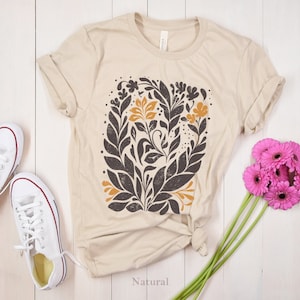 May include: A beige t-shirt with a floral design in black, brown, and yellow. The design features leaves and flowers. The shirt is laid flat on a white wooden surface. The word "Natural" is printed on the surface below the shirt.