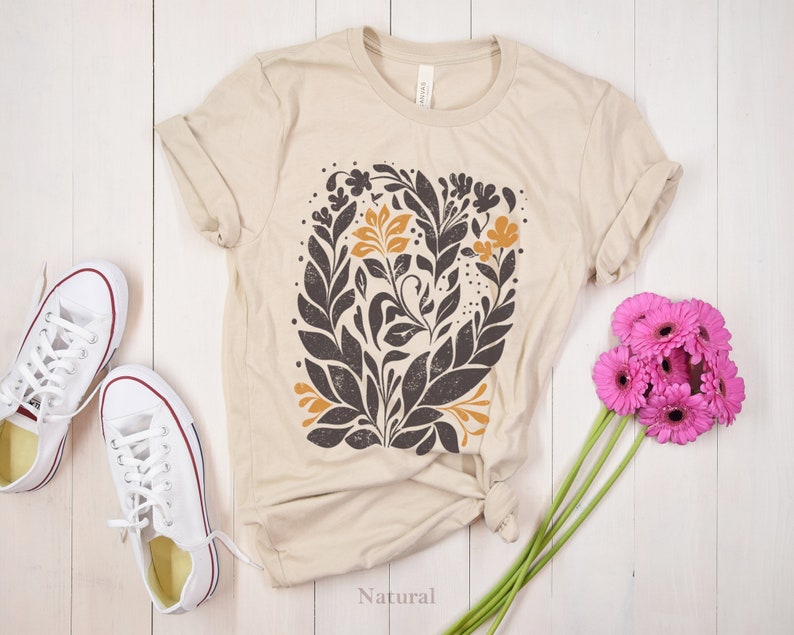 May include: A beige t-shirt with a floral design in black, brown, and yellow. The design features leaves and flowers. The shirt is laid flat on a white wooden surface. The word "Natural" is printed on the surface below the shirt.