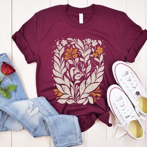May include: A maroon t-shirt with a white and orange floral design. The shirt is laid out on a white wooden surface with a pair of white sneakers and blue jeans.