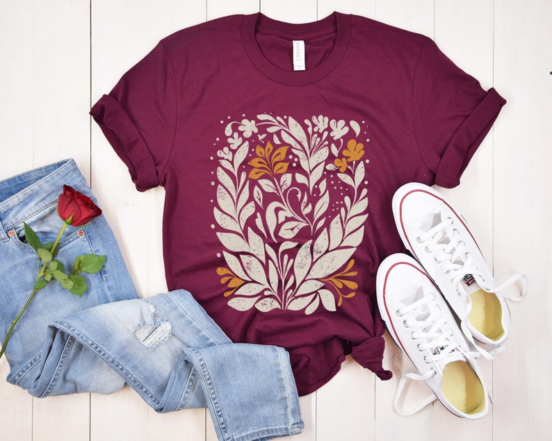 May include: A maroon t-shirt with a white and orange floral design. The shirt is laid out on a white wooden surface with a pair of white sneakers and blue jeans.