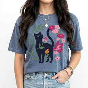 Pu&ograve; includere: Una t-shirt blu a maniche corte con la grafica di un gatto nero circondato da foglie verdi e fiori rosa e rossi. Il gatto &egrave; raffigurato di profilo con una lunga coda rivolta verso l'alto e il disegno include un piccolo elemento botanico arancione.