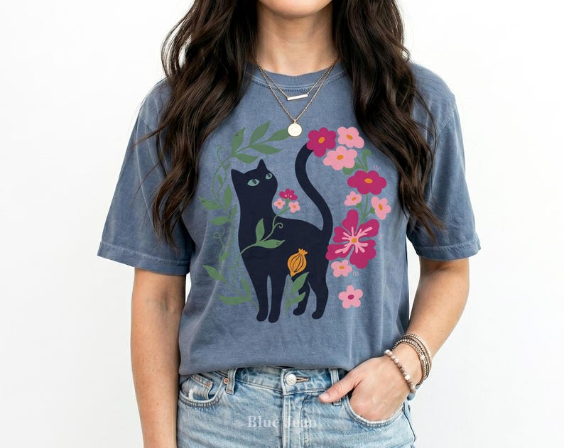 Pu&ograve; includere: Una t-shirt blu a maniche corte con la grafica di un gatto nero circondato da foglie verdi e fiori rosa e rossi. Il gatto &egrave; raffigurato di profilo con una lunga coda rivolta verso l'alto e il disegno include un piccolo elemento botanico arancione.