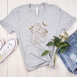 K&ouml;nnte beinhalten: Hellgraues T-Shirt mit goldener Strichzeichnung einer Frau, einem Mond und floralen Akzenten. Das Shirt wird mit Jeansshorts und wei&szlig;en Turnschuhen kombiniert.