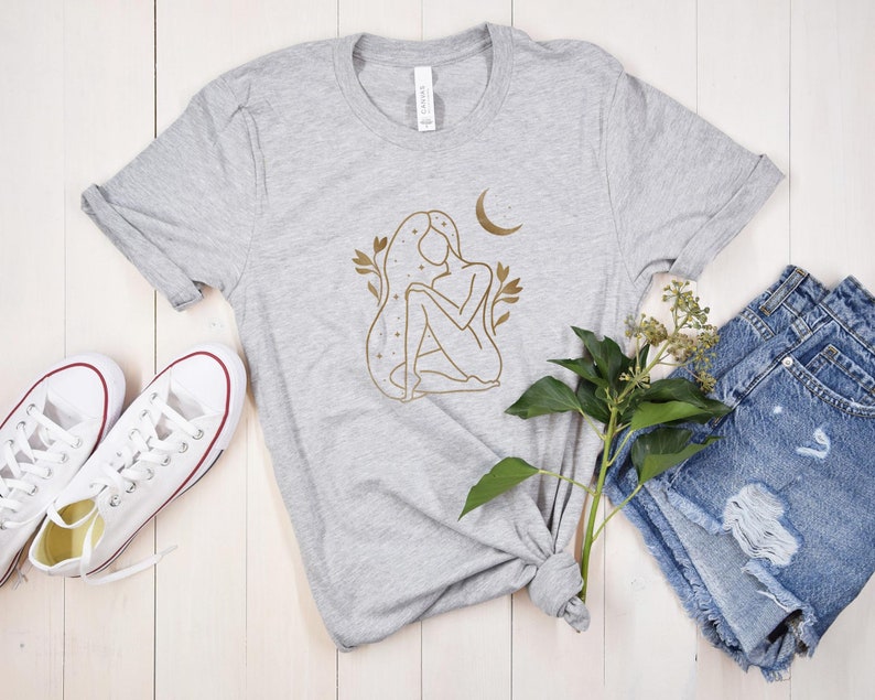 K&ouml;nnte beinhalten: Hellgraues T-Shirt mit goldener Strichzeichnung einer Frau, einem Mond und floralen Akzenten. Das Shirt wird mit Jeansshorts und wei&szlig;en Turnschuhen kombiniert.