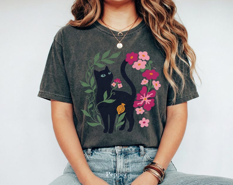 Pu&ograve; includere: Una t-shirt a maniche corte color grigio antracite con una stampa grafica di un gatto nero circondato da viti verdi e fiori rosa e magenta. Il gatto &egrave; posizionato centralmente con la coda curva verso l'alto.