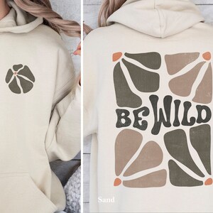 Op de afbeelding: Een beige hoodie met een bloemetjesontwerp aan de voorkant en de woorden "BE WILD" in een retro-lettertype aan de achterkant. Het ontwerp aan de achterkant heeft een geometrisch patroon in bruine en groene tinten.