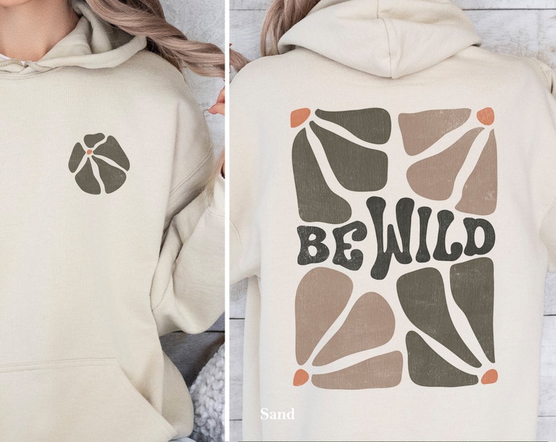 Op de afbeelding: Een beige hoodie met een bloemetjesontwerp aan de voorkant en de woorden "BE WILD" in een retro-lettertype aan de achterkant. Het ontwerp aan de achterkant heeft een geometrisch patroon in bruine en groene tinten.