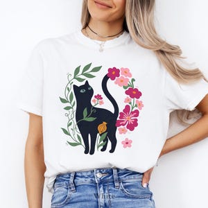 Pu&ograve; includere: Una t-shirt bianca con un'illustrazione grafica di un gatto nero circondato da viti verdi e vari fiori rosa e rossi. Il gatto &egrave; raffigurato di profilo, rivolto verso l'alto, con la coda elegantemente curva tra gli elementi floreali.