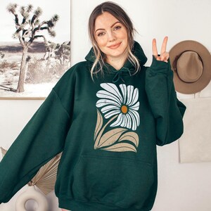 K&ouml;nnte beinhalten: Ein dunkelgr&uuml;ner Kapuzenpullover mit einem wei&szlig;en G&auml;nsebl&uuml;mchen-Druck. Die G&auml;nsebl&uuml;mchen haben eine gelbe Mitte und braune Bl&auml;tter.