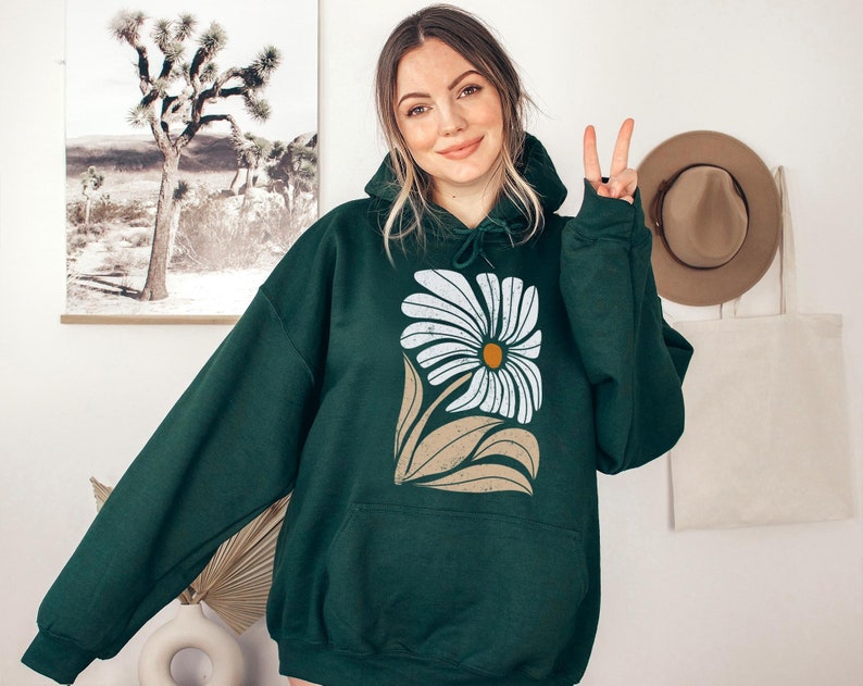 K&ouml;nnte beinhalten: Ein dunkelgr&uuml;ner Kapuzenpullover mit einem wei&szlig;en G&auml;nsebl&uuml;mchen-Druck. Die G&auml;nsebl&uuml;mchen haben eine gelbe Mitte und braune Bl&auml;tter.