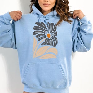 K&ouml;nnte beinhalten: Ein hellblauer Hoodie mit einem stilisierten Blumendesign in Schwarz, Orange und Beige. Die Blume hat eine gro&szlig;e, dunkle Mitte und Bl&uuml;tenbl&auml;tter, die gebogen und spitz sind.