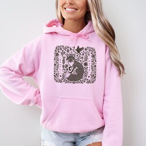 May include: Pale pink hooded sweatshirt with a front pocket. The design features a fox surrounded by floral elements and a bird. The graphic is in shades of brown and black.