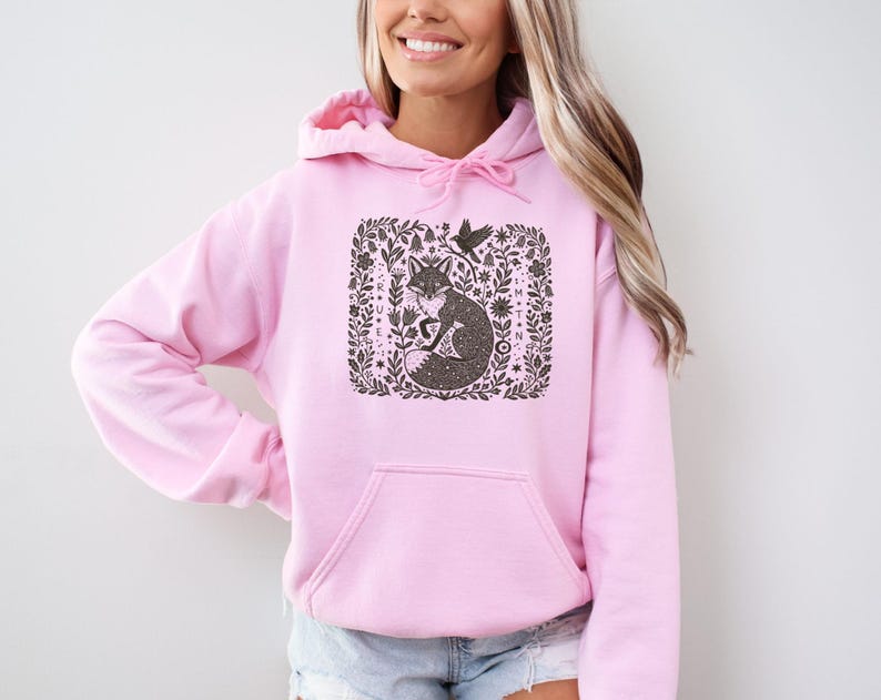 May include: Pale pink hooded sweatshirt with a front pocket. The design features a fox surrounded by floral elements and a bird. The graphic is in shades of brown and black.
