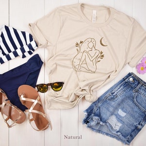 K&ouml;nnte beinhalten: Beiges T-Shirt mit goldener Illustration einer Frau, einem Mond und floralen Elementen. Das Bild zeigt auch ein gestreiftes Bikini-Oberteil, eine marineblaue Bikinihose, Jeansshorts, goldene Sandalen und eine Sonnenbrille. Das Wort "Natural" ist unten aufgedruckt.