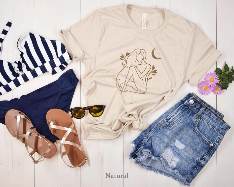 K&ouml;nnte beinhalten: Beiges T-Shirt mit goldener Illustration einer Frau, einem Mond und floralen Elementen. Das Bild zeigt auch ein gestreiftes Bikini-Oberteil, eine marineblaue Bikinihose, Jeansshorts, goldene Sandalen und eine Sonnenbrille. Das Wort "Natural" ist unten aufgedruckt.