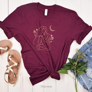 K&ouml;nnte beinhalten: Burgunderfarbenes T-Shirt mit goldenem Linienkunst-Design einer Frau, eines Mondes und Sterne. Das Shirt wird mit goldenen Sandalen und Jeansshorts gestylt. Das Wort "Maroon" ist unten auf dem Shirt aufgedruckt.