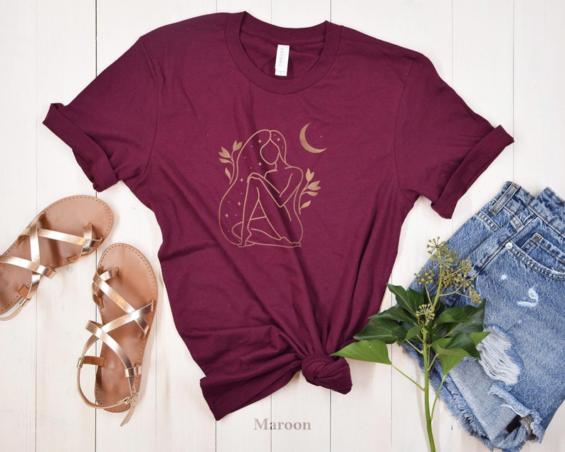 K&ouml;nnte beinhalten: Burgunderfarbenes T-Shirt mit goldenem Linienkunst-Design einer Frau, eines Mondes und Sterne. Das Shirt wird mit goldenen Sandalen und Jeansshorts gestylt. Das Wort "Maroon" ist unten auf dem Shirt aufgedruckt.