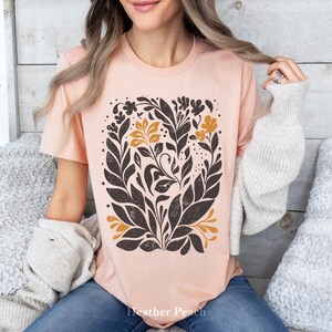 May include: A peach-colored t-shirt with a black and gold floral design. The design features stylized leaves and flowers in a repeating pattern. The text "Heather Peach" is printed on the bottom of the shirt.