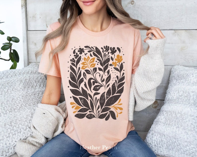May include: A peach-colored t-shirt with a black and gold floral design. The design features stylized leaves and flowers in a repeating pattern. The text "Heather Peach" is printed on the bottom of the shirt.