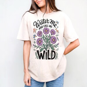 May include: A cream colored t-shirt with a floral design and the text "Water Me And Call Me Wild".