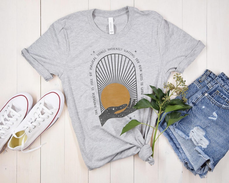 May include: A gray t-shirt with a graphic design of a hand holding a sun with the text "The universe is full of magical things patiently waiting for our wits to grow".