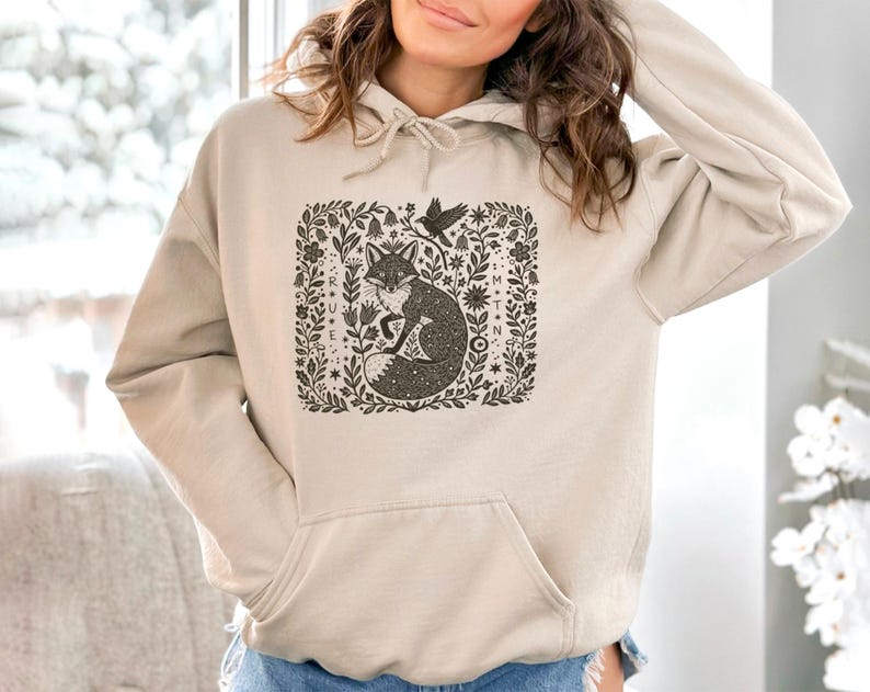 May include: Beige hoodie with a front pocket and a drawstring hood. The graphic on the front features a detailed illustration of a fox, a bird, and floral elements in a square frame. The text "TRUE MOON" is also visible.