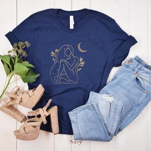 K&ouml;nnte beinhalten: Marineblaues T-Shirt mit goldener Illustration einer Frau, Mond und Sternen. Das Shirt wird mit hellblauen Jeans und beigen Plateausandalen kombiniert. Das Wort "Navy" ist unten im Bild aufgedruckt.