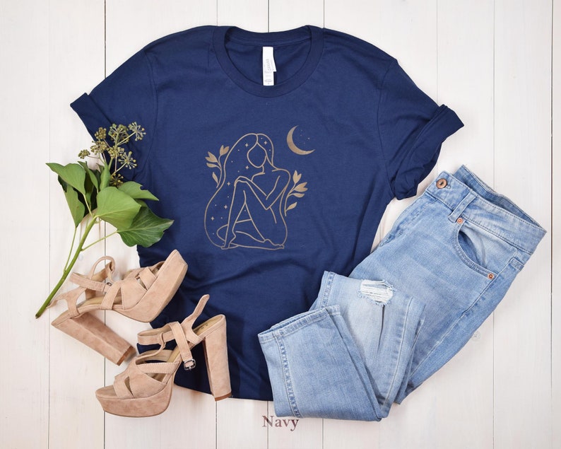 K&ouml;nnte beinhalten: Marineblaues T-Shirt mit goldener Illustration einer Frau, Mond und Sternen. Das Shirt wird mit hellblauen Jeans und beigen Plateausandalen kombiniert. Das Wort "Navy" ist unten im Bild aufgedruckt.