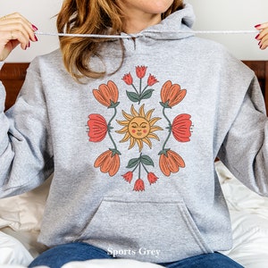 May include: A gray hooded sweatshirt with a graphic print of a sun with a face and flowers in shades of orange, red, and green. The text "Sports Grey" is printed on the bottom of the sweatshirt.